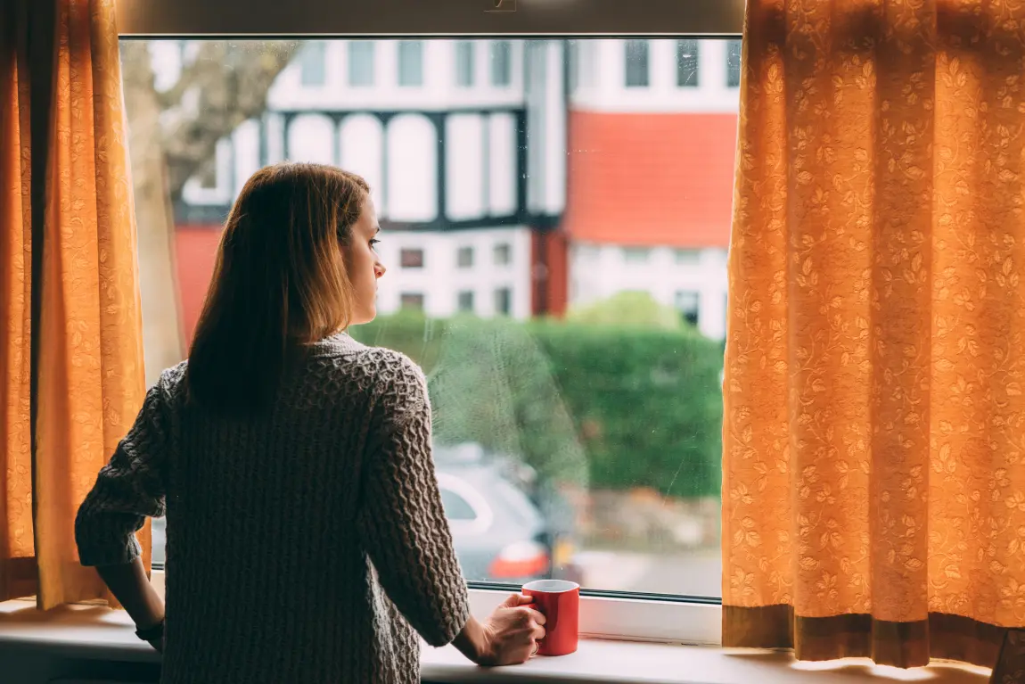 Kvinna som står och tittar ut genom ett fönster med en mugg i handen